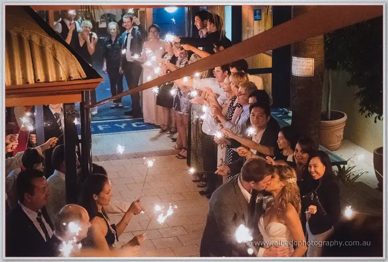 Bride and groom kiss beneath a tunnel of guests waving sparklers outside the yacht club at night