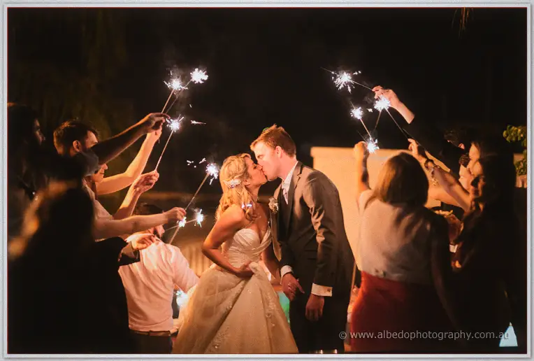 Newlyweds kiss ringed by guests lifting sparkling sparklers overhead into the night air
