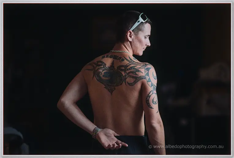 A shirtless groomsman with a full back tattoo poses bare-chested while getting ready indoors.