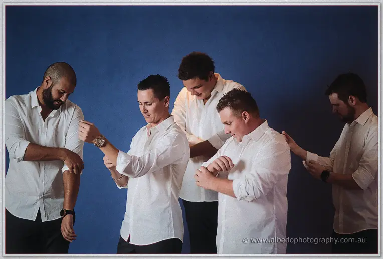 Five groomsmen in white shirts roll their sleeves in unison against a deep blue wall.