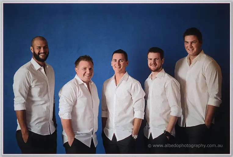 Five groomsmen in crisp white shirts grin together against a deep blue wall before the ceremony.