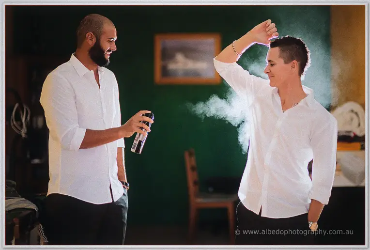 A groomsman sprays deodorant while the groom laughs and flexes playfully in the green room.