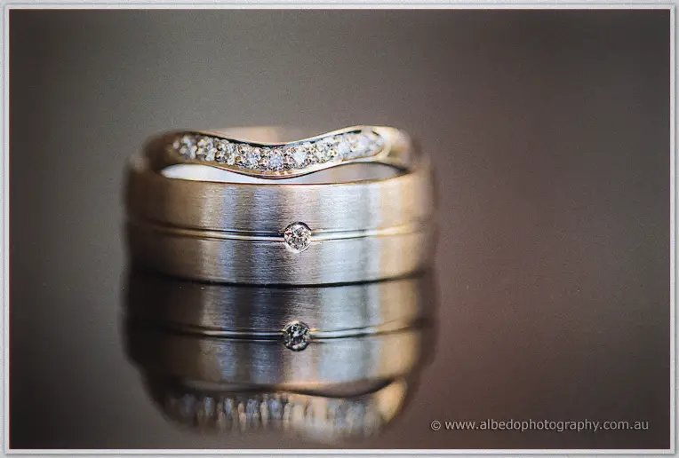 Elisha and Shane's wedding bands stack on a reflective surface, a diamond-set wave band atop his ring.