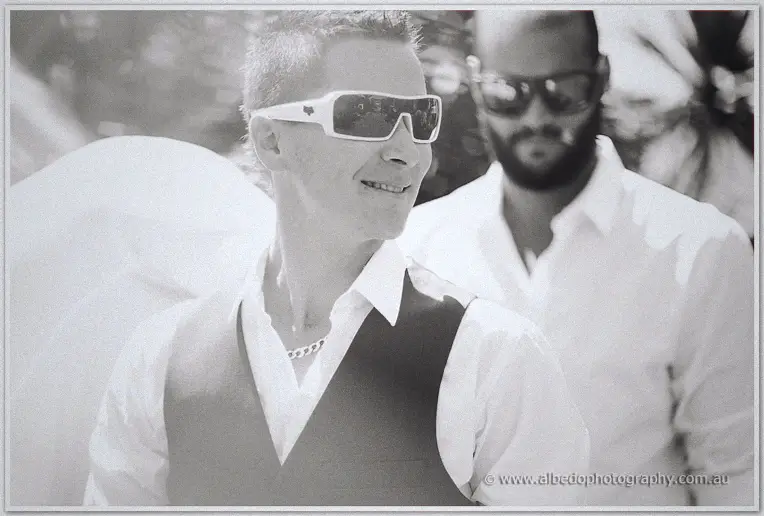 In black and white, Shane smiles in white sunglasses while a groomsman follows with his white shirt.
