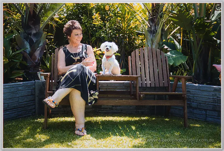 A smiling guest in a black dress shares a garden bench with the couple's white maltese terrier.