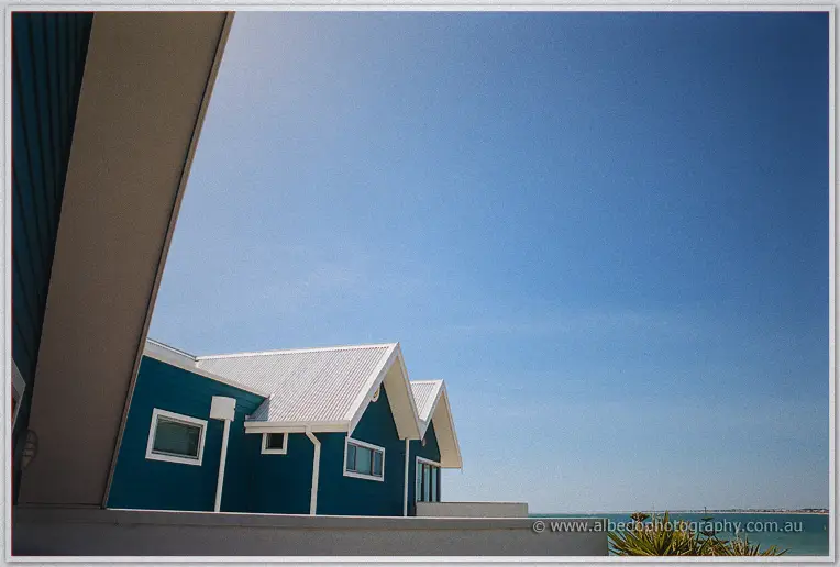 White gables of the beach house rise against a clear sky, turquoise water glimpsed beyond the balcony.