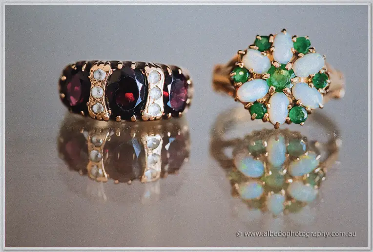 Two vintage keepsake rings, garnet-and-pearl and opal-and-emerald, gleam side by side on glass.