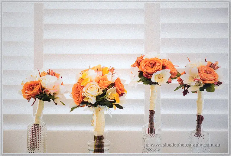 Four bouquets of orange roses and cream blooms wait in glass vases against the white shutters.