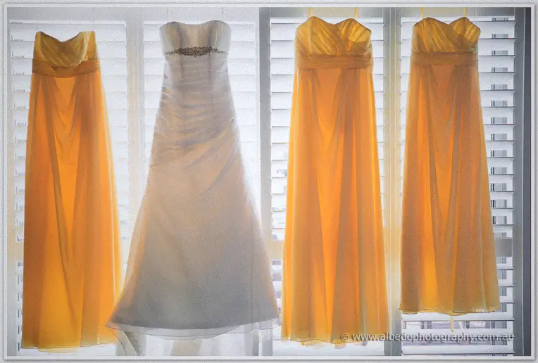 Elisha's white gown hangs between three tangerine bridesmaid dresses, backlit by the shutters.