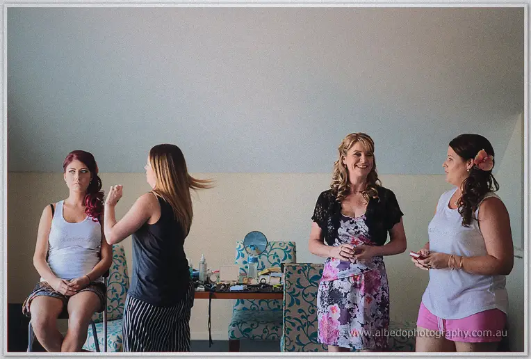 A stylist curls a bridesmaid's hair as two women chat over makeup in the sunlit getting-ready room.