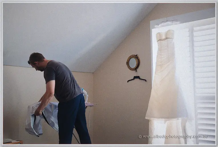 A groomsman irons his shirt beside the window where Elisha's gown waits in the soft morning light.