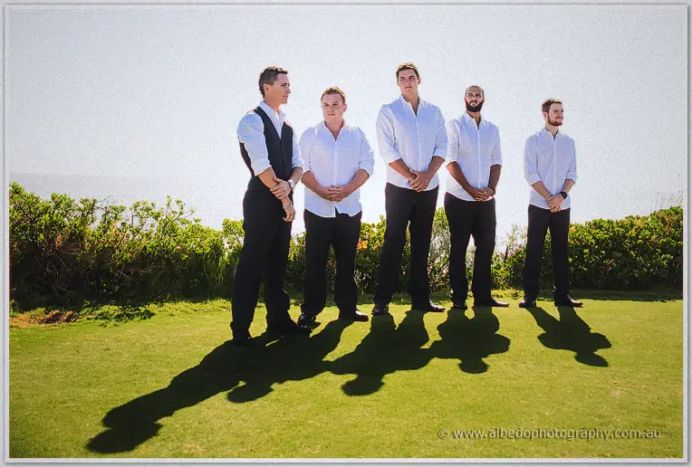 Shane and his groomsmen stand in a line on the green, casting long shadows by the coast