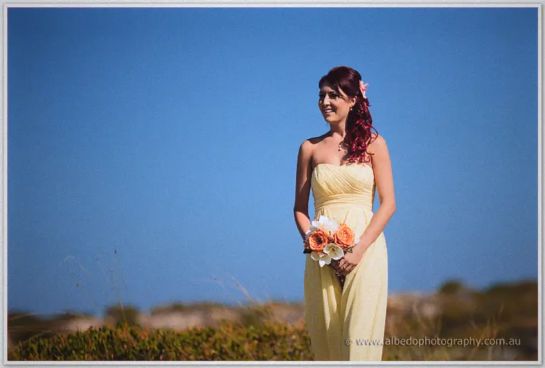 A dark-haired bridesmaid smiles holding peach roses against a blue sky
