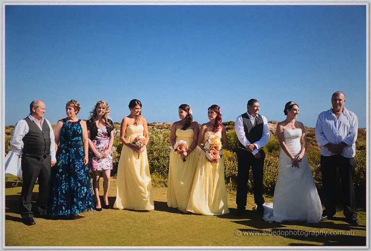 Family and bridesmaids in yellow line up beside the couple during the clifftop ceremony