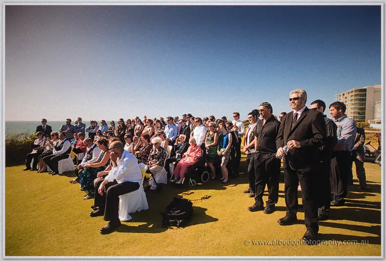 Wedding guests seated and standing across the lawn with the ocean behind them