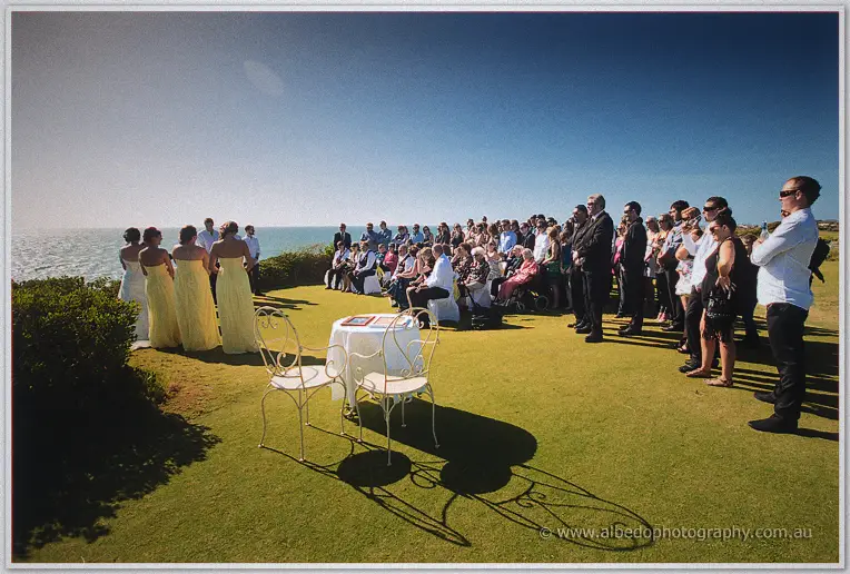 Wide view of the clifftop ceremony with guests facing the ocean and ornate white chairs
