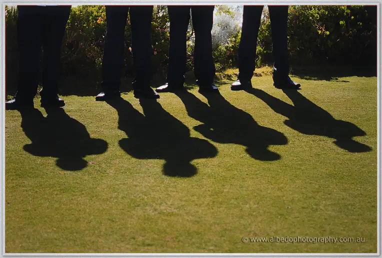 Low shot of groomsmen's legs and long shadows cast on the sunlit lawn