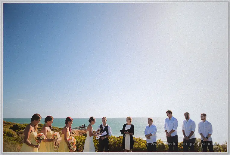 Wide ceremony scene with celebrant, couple, and party set against the sea horizon