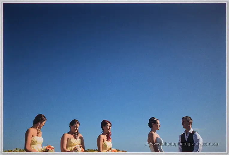 Bridesmaids and the couple framed small beneath a vast blue sky at the clifftop
