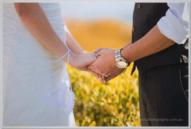 The couple's joined hands and ring pillow glow in golden coastal light