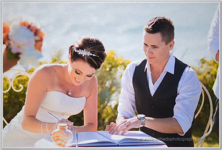 Elisha signs the marriage register as Shane rests his hand beside hers by the sea