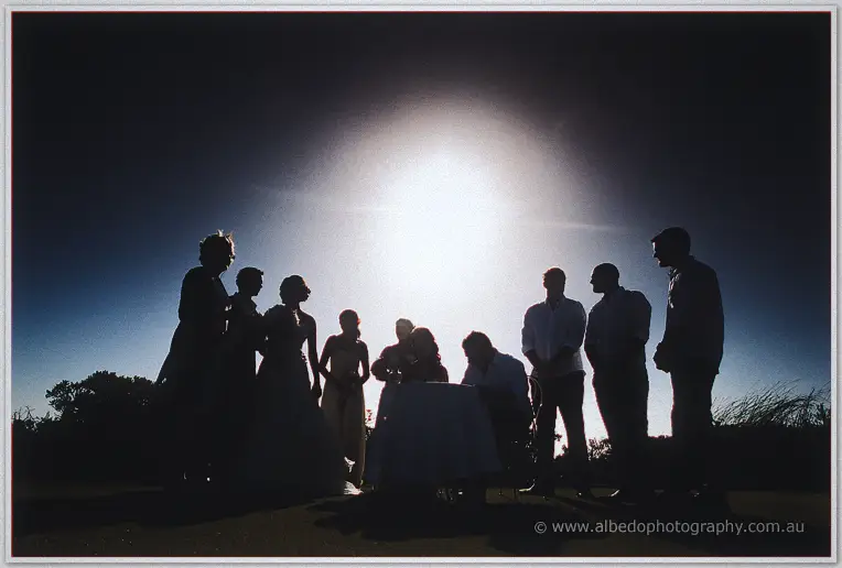 Silhouettes of the couple and guests circle the signing table against the low sun