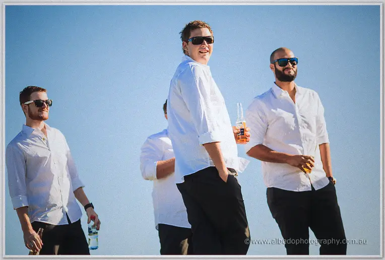 Groomsmen in white shirts and sunglasses relax with beers under the afternoon sun