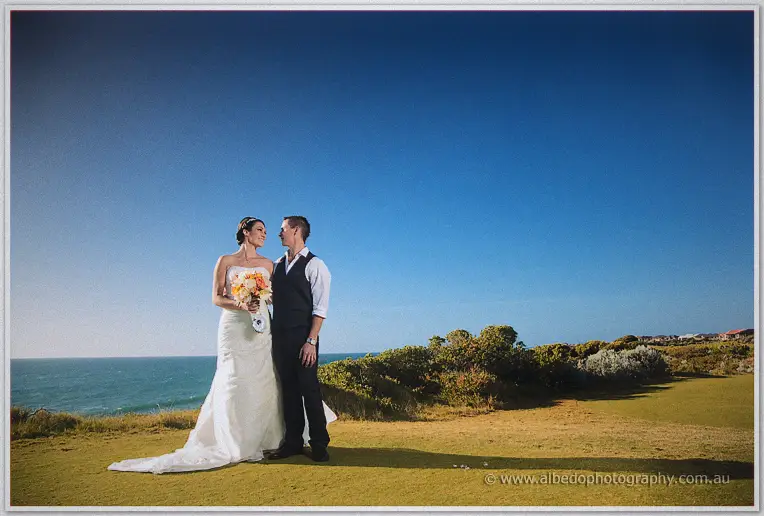 Elisha and Shane stand close on the clifftop green with the ocean sweeping behind