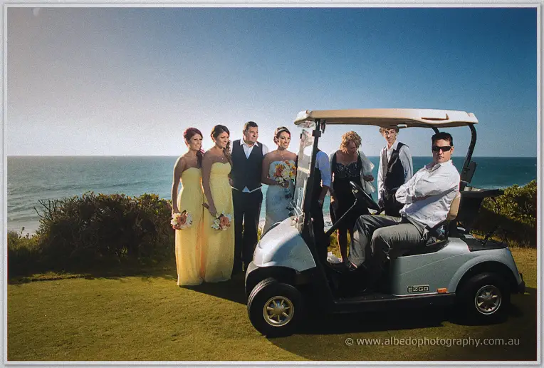 Bridal party leans around a golf cart with the sea stretched out behind them