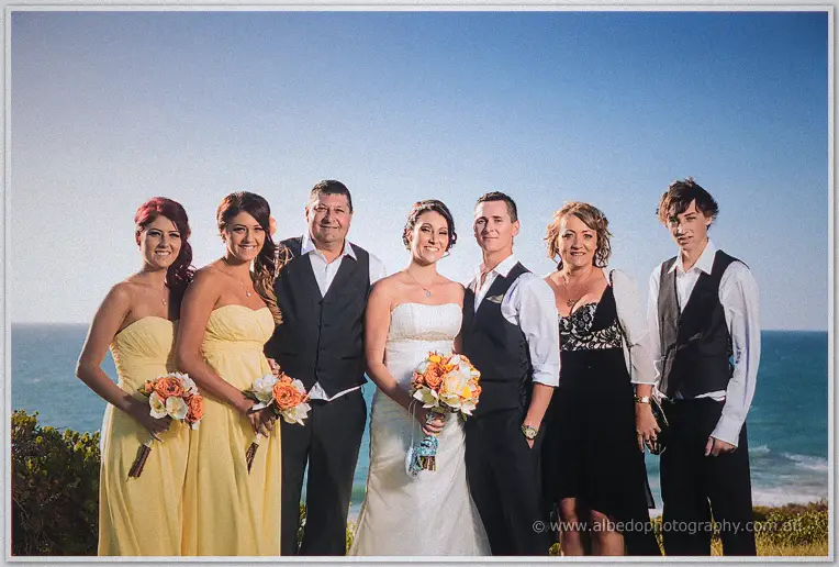 Elisha and Shane pose with family holding peach bouquets above the coastline