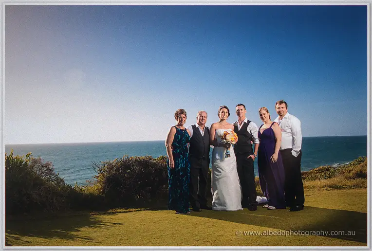 Six family members in blue and purple flank the couple on the clifftop lawn