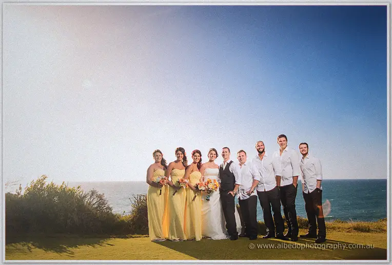 Bridal party in yellow gowns and white shirts gathered on a coastal green above the ocean at The Cut