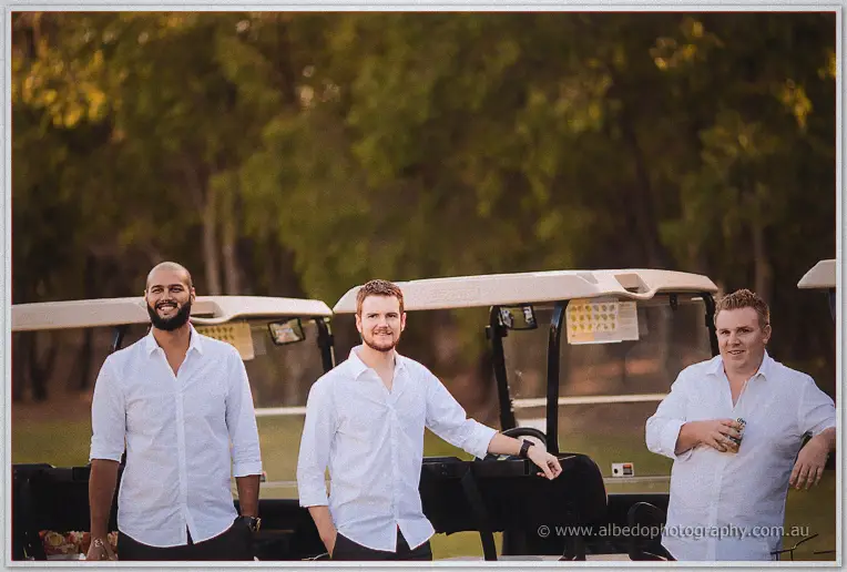 Groomsmen in white shirts leaning on golf carts among the trees, sharing a relaxed pre-ceremony moment