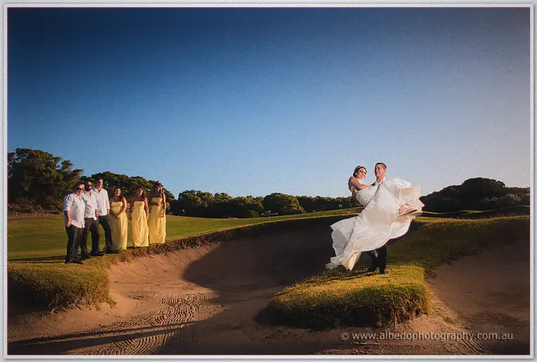 Groom carries his laughing bride across a sand bunker as the wedding party watches from the green