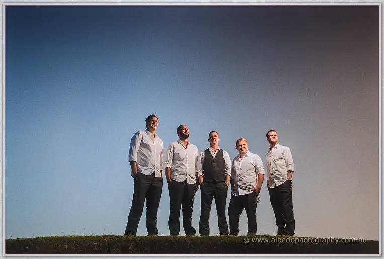 Five groomsmen stand shoulder to shoulder on a ridge against a clear sky, shot from below