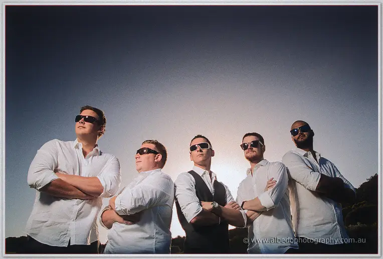 Groomsmen in sunglasses and folded arms lined up against a hazy backlit sky at The Cut