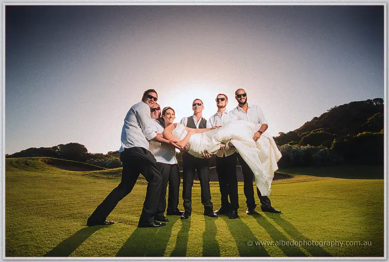 Playful group lifts the bride in her gown on the fairway, sun flaring behind the laughing party