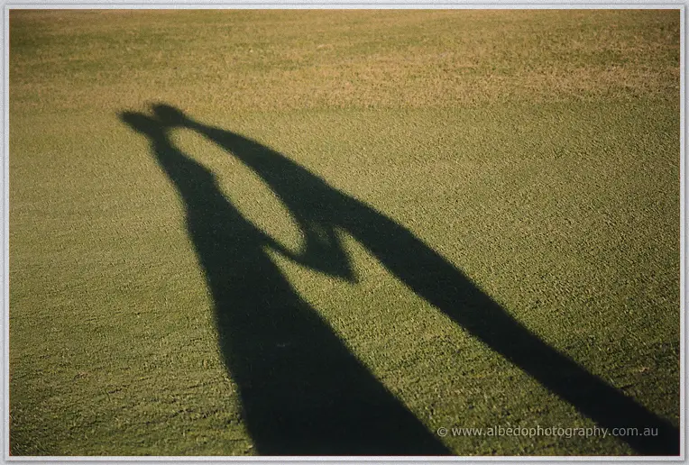 Long shadows of the newlyweds holding hands stretch across the mown green in low sun