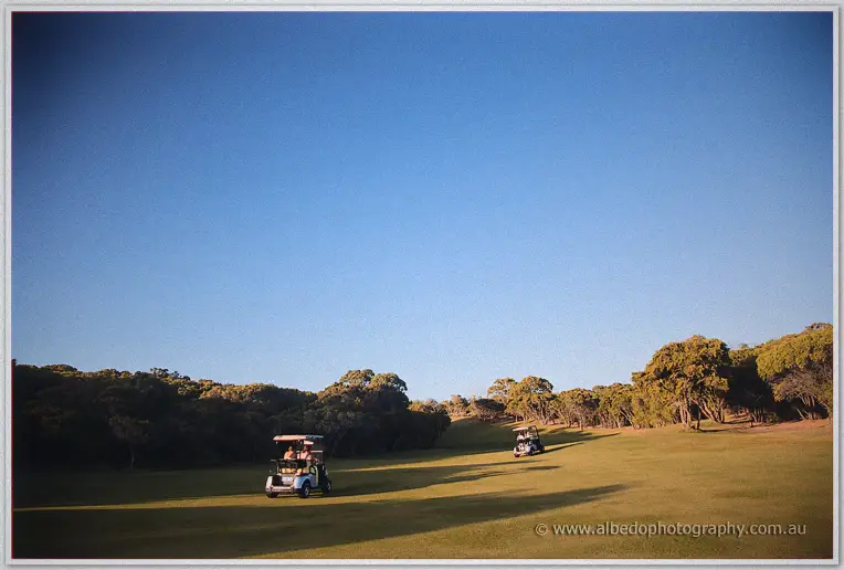 Two golf carts carry the wedding party down a wide sunlit fairway under a clear sky