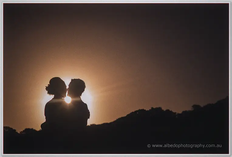 Silhouetted newlyweds kiss against the glowing sun as it sinks behind the treeline