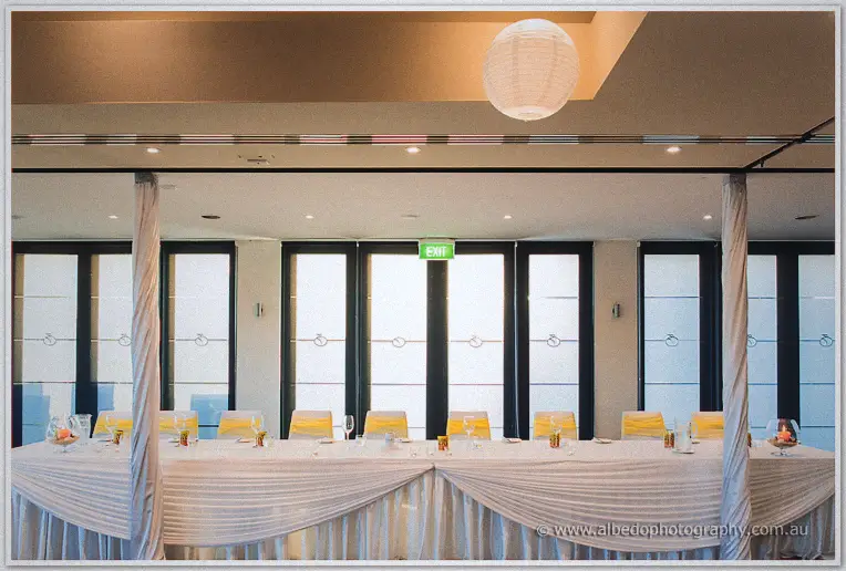 Long bridal table dressed in white with yellow sashes waits under paper lanterns