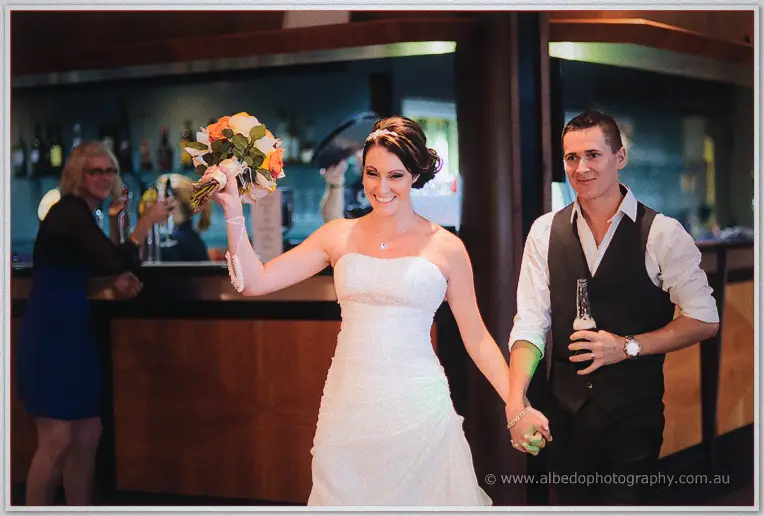 Elisha raises her bouquet while leading Shane by the hand into the reception past the bar