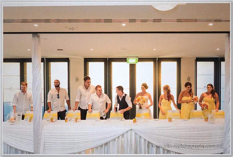 Full bridal party lines the long table at the reception, groomsmen in white and maids in yellow
