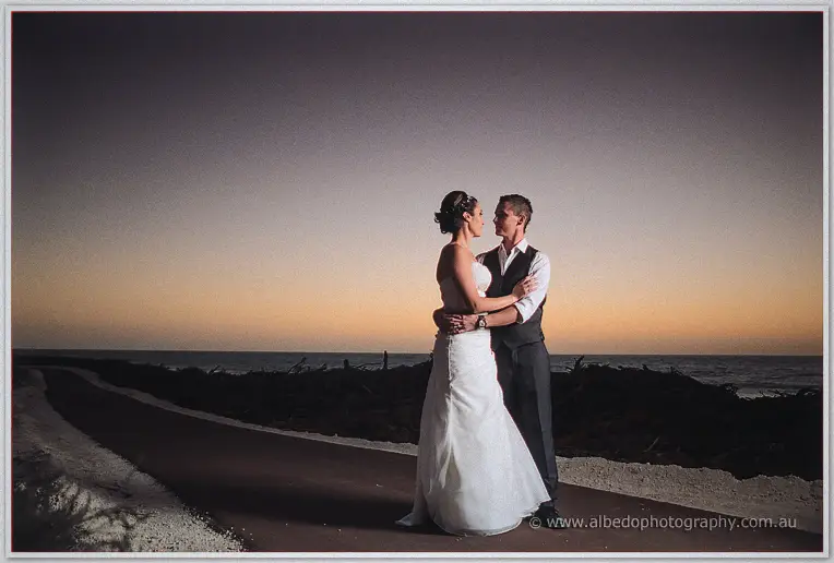Newlyweds embrace on a coastal path at twilight with the last orange glow behind them
