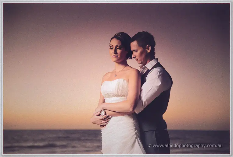 Shane wraps his arms around Elisha on the sand under a soft pink and amber dusk