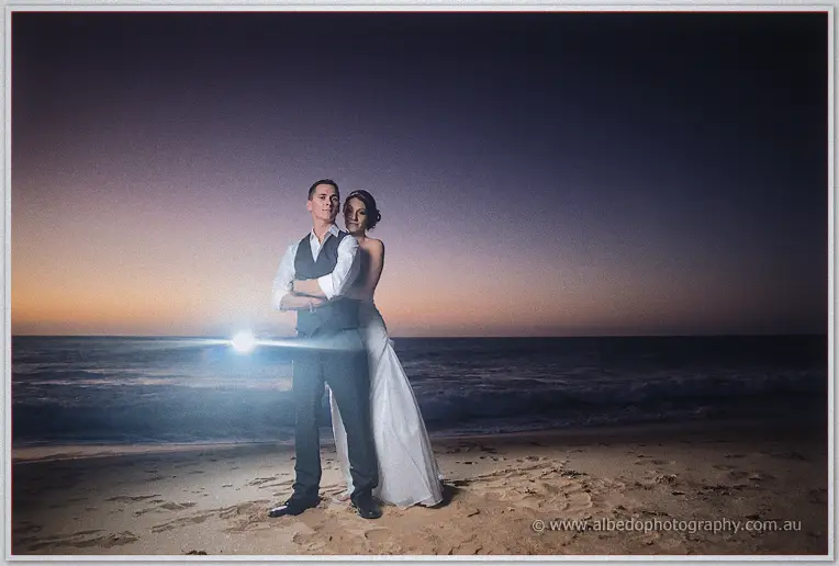 Couple stands backlit on the beach at twilight, flare from an off-camera light behind them