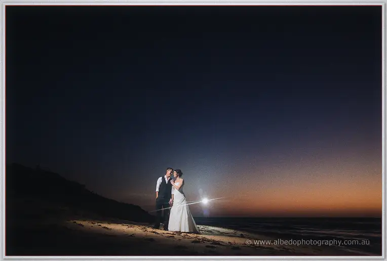 Wide night scene of the newlyweds alone on the dark beach beneath a fading sunset glow