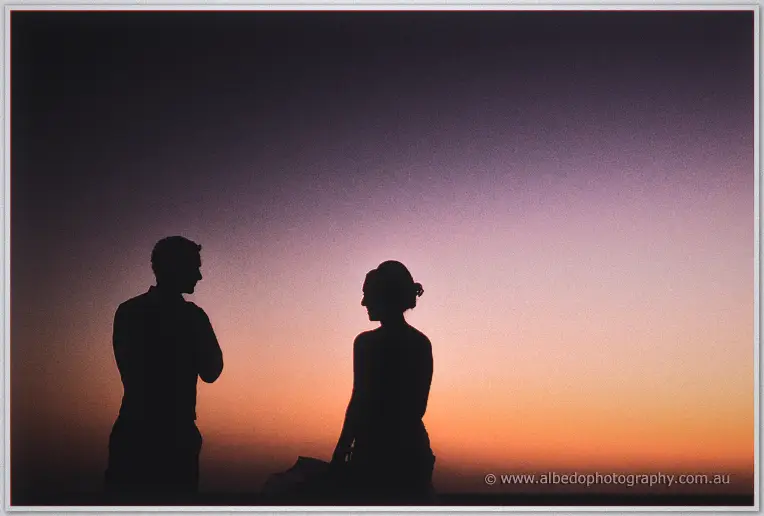Bride and groom stand as black silhouettes against a glowing orange and mauve sunset