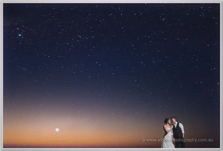 The couple embrace beneath a vast starry sky with the moon low over the glowing horizon