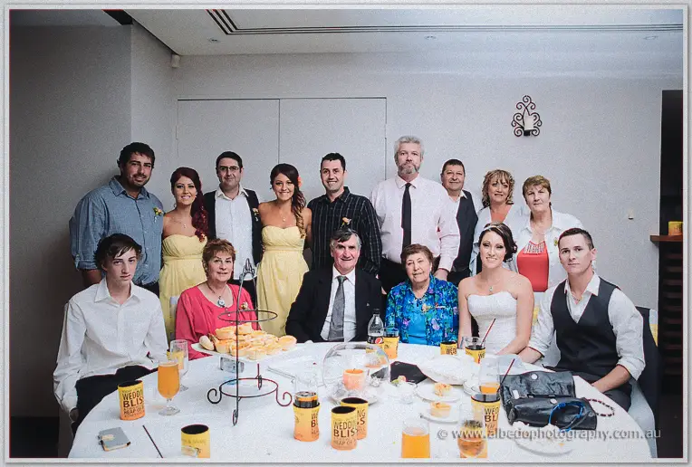 Family and friends crowd a round table with the couple for a group photo indoors